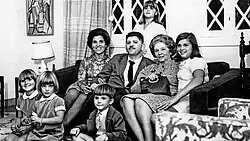 Sur un canapé, dans un salon, un homme en costume entouré de 4 femmes dont deux jeunes femmes. Devant, assis par terre trois enfants.