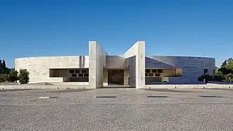 Photographie couleur d’un édifice moderne couleur béton donnant sur un grand parvis sec.