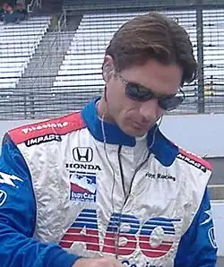 Photographie d'un homme, vu de trois-quarts, en gros plan, avec des lunettes de soleil, la tête vers le bas.
