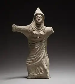 petite statuette de femme vue de face, portant un chapeau pointu, les seins dénudés, le reste du corps drapé dans un long vêtement, bras ouverts sur le côté