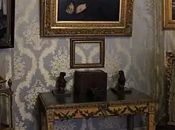 Photo d'intérieur d'un musée, agrandie sur une fine table comportant quelques objets. Au-dessus, un cadre vide laisse apparaître la tapisserie bleue du mur.