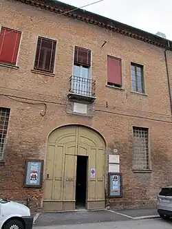 Façade de maison en brique rouge avec à l'étage Quatre fenêtres multicolores, avec à son centre un grand portail vert clair à arc de cercle avec de chaque côté une fenêtre à barreaux, surmonté d'une fenêtre terrasse