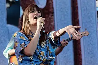Femme jeune avec les cheveux longs châtains, photographiée de face, en train de chanter, bras gauche; le manche d'une basse et la main du bassiste sont visibles dans l'arrière-plan à droite.