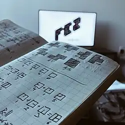 Photographie d'un carnet de notes sur lequel quelqu'un a grisé des carreaux pour reproduire les caractères mystérieux qu'affiche le jeu dans ses énigmes.