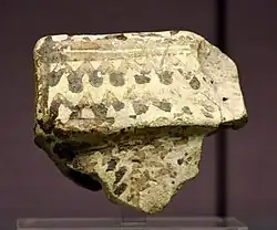 Morceau de terre cuite beige avec quelques traits de peinture noire.