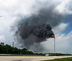 Vue au niveau du sol d'une haute torchère émettant un nuage de fumée noire, au milieu d'un ciel bleu. Au premier plan figure une route et des arbres sont présents à gauche de l'image.