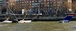 Photo montrant l'avion au premier plan amarré à une jetée, partiellement submergé dans le fleuve Hudson. Il est relié par des câbles à la berge et seule sa queue et son aile gauche sont clairement visibles, le reste de son corps se trouvant sous l'eau.