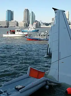 Photo de l'avion amarrée sur la rive. La photo, prise depuis l'intérieur de l'avion, montre l'aile et un toboggan d'évacuation ainsi que la ville de New York à l'arrière-plan.