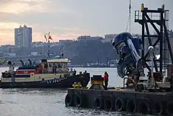 Photo de la récupération du moteur. À gauche, un navire supervise les opérations tandis qu'à droite, sur la berge, une grue tire le moteur endommagé du fleuve.