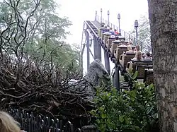 Un manège de montagnes russes s'élève au-dessus des végétations. À gauche, on aperçoit la tête d'aigle grise d'une créature factice.