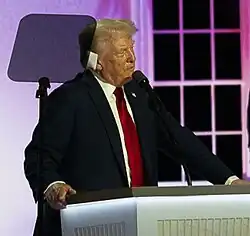 Homme en costard devant un microphone avec un pansement sur l'oreille.