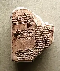 Photographie d'une tablette  fragmentée en argile beige couverte d'écritures cunéiformes anciennes. La tablette est inégale et incomplète, avec des bords cassés. Elle montre des signes d'érosion qui témoignent de son âge et de son origine historique. Les lignes de texte cunéiforme sont incomplètes.