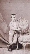 Photographie ancienne d'un petit garçon en costume, accoudé à un fauteuil.