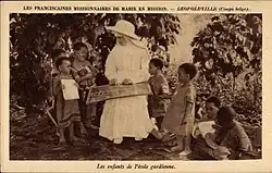 7 bambins debout ou assis au sol, de part et d'autre d'une religieuse assise, souriante, vêtue de blanc et chapeautée, leur montrant du doigt le détail d'un document placé sur ses genoux.