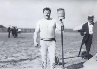 Portrait d'un homme à moustache portant une tunique claire et tenant en main une barre surmontée de poids