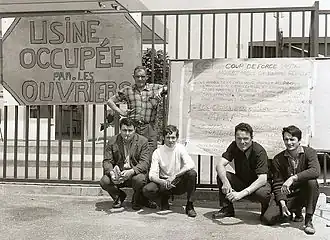 Photo en noir et blanc de cinq hommes accroupis ou debout devant une grille où figurent un écriteau « Usine occupée par les ouvriers » et un panneau d'information syndicale portant le sigle de la CGT