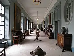 Longue galerie avec fenêtre à gauche et portes à droite, ponctuée de meubles et de tableaux