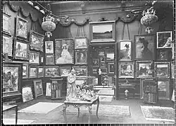 Vue d'une partie d'une salle de la galerie Henry Vasnier. Sur les murs sont accrochés des tableaux, d'autres sont posés à même le sol, contre eux. Au centre se trouve une table sur laquelle sont posés des sculptures et des objets d'art. Des livres sont rangés dans des meubles devant les cimaises. Des tapis recouvrent le sol. Une porte ouverte laisse entre voir la salle suivante.