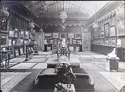 Vue d'ensemble d'une salle de la Galerie Vasnier avec la verrière comme source de lumière zénithale. Les murs sont recouverts de tableaux. Devant les cimaises, les sculptures sont disposées sur des sellettes et les objets d'art dans une vitrine. Des tapis recouvrent le sol.
