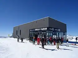 Photographie d'une gare de téléphérique avec des skieurs, en tôles grises et sur un socle montagneux enneigé.