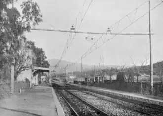 Photographie en noir et blanc d'une gare de taille moyenne avec voies électrifiées.