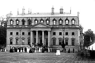 Photographie ancienne en noir et blanc d'un manoir néoclassique devant lequel une foule est réunie pour un évènement festif.