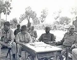 Photographie en noir et blanc montrant quatre hommes, dont deux en uniforme, assis autour d'une table dans un cadre naturel. D'autres personnes sont présentes en arrière-plan.