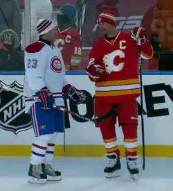 Photographie de deux joueurs de hockey sur glace discutant. L'un porte un uniforme blanc, bleu et rouge, l'autre rouge, blanc et jaune.