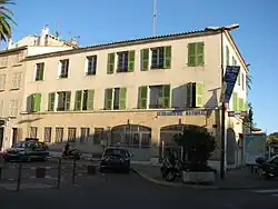 Un long bâtiment de style provençal avec des barreaux aux ouvertures du rez-de-chaussée et des volets verts aux deux étages.