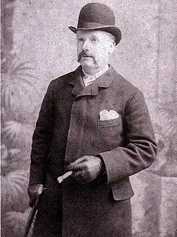 Photographie en noir et blanc. Homme au chapeau melon, avec manteau et gants, une canne sous le bras droit.