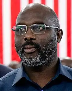 Portrait d'un homme noir chauve et portant une barbe poivre et sel. Il porte une chemise bleu marine et des lunettes.