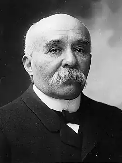 Photo d’un homme âgé, cheveux blancs, calvitie prononcée, épaisse moustache blanche, portant un nœud papillon, une chemise blanche et une veste noire