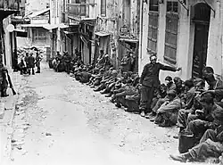 photographie noir et blanc : des hommes assis le long de maisons dans une rue en pente