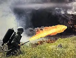 Photo en couleur montrant un soldat de dos utilisant un lance-flammes. Une grande flamme jaune jaillie du tuyau et traverse l'image, l'arrière plan est totalement obstrué par une épaisse fumée noire.