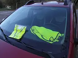 Photographie du pare-brise d'une voiture à l'arrêt, vue de l'extérieur. Deux gilets jaunes sont exposés de façon à ce qu'ils soient très visibles.