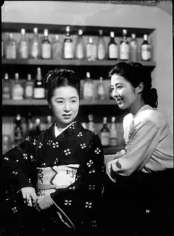 Une femme en kimono sombre (Kinuyo Tanaka) se tient de face au comptoir d'un bar. À droite, une jeune femme en chemisier blanc (Kyōko Kagawa) la regarde en souriant. Dans le fond, plusieurs étagères supportent des rangées de bouteilles (image en noir et blanc).