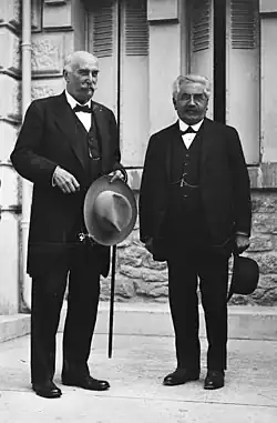 Deux hommes moustachus et vêtus chacun d’un costume foncé sourient sur le perron d'un bâtiment : à gauche, l’homme est chauve, tient dans sa main gauche un chapeau clair et une canne ; à droite, l'individu est plus petit, a les cheveux gris-blancs et tient dans sa main gauche un chapeau de couleur foncée