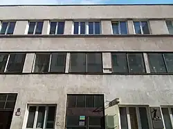 Photographie d'un bâtiment à la façade beige, aux fenêtres rectangulaires ; en bas à droite de l'image, une plaque avec un texte et une représentation artistique.