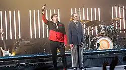 Photo de deux hommes blancs et bruns debout sur une scène devant des instruments de musique. Celui de gauche porte une chemise rouge et noire et un pantalon noir. Celui de droite a de petites lunettes rondes, un costume-cravate et une petite moustache.