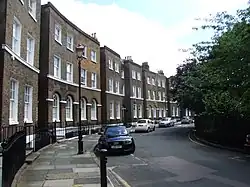 Rue pavée étroite bordée de maisons en briques brunes à étages. Voitures garées, arbres verdâtres, lampadaire central. Perspective horizontale, atmosphère calme de quartier résidentiel.