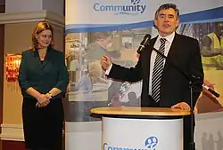 Sur la droite, un homme souriant aux cheveux courts et en costume-cravate s'exprime au micro devant un pupitre en levant sa main droite. Sur la gauche, une femme aux cheveux roux mi-longs portant un tailleur le regarde en souriant dans une attitude posée, joignant ses mains devant elle