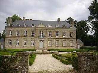 Photographie en couleur montrant la façade d'un château néo-classique, devant une cour avec une fontaine entourée de haies taillées.