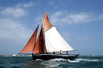 Photographie d’un sloop sardinier, voiles oranges et blanche sur fond de ciel bleu.
