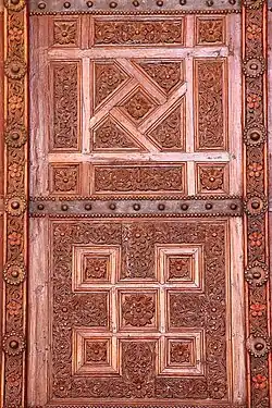 Photographie d'un vantail en bois sculpté de la grande porte de la salle de prière. Des bandes de tôle, ornées de clous à tête ronde, séparent les panneaux ouvragés de chaque vantail.