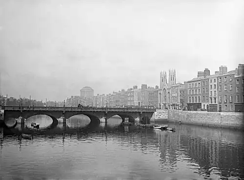 Photographie ancienne, en noir et blanc, d'un pont aux arches de pierre traversant un fleuve au milieu d'une ville.