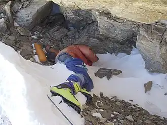 Photographie couleur en plongée d'un homme allongé entre neige et roche et au visage caché derrière son bras, emmitouflé dans d'épais vêtements et portant des chaussures d'alpinisme vertes.