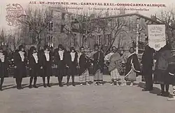 Carte postale noir et blanc d'un groupe d'une douzaine de personnes debout. Un bâtiment an arrière-plan.