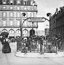 Photo noir et blanc d'une place avec une balustrade et de hauts candélabres autour d'un escalier ; devant à gauche, une femme en jupe longue ; derrière, un grand immeuble