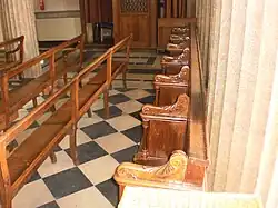 Bois sculpté et vernis, le siège est adossé à un mur et une colonne, et présente plusieurs accoudoirs.