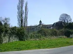 Derrière une végétation sauvage, on aperçoit la lanterne de la chapelle, les étages du bâtiment principal et, à droite, le pavillon nord. Au premier plan, brèche dans le mur d'enceinte, que colmate un grillage en mauvais état.